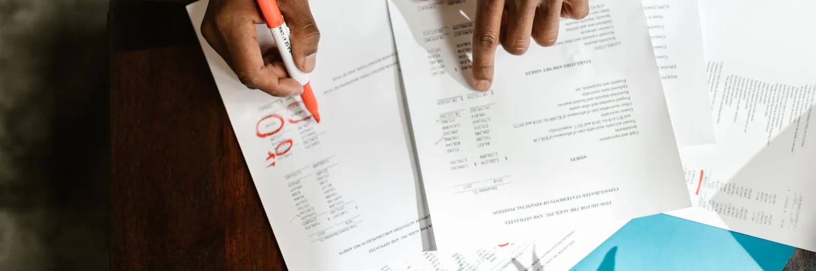Overhead flat-lay of two hands marking up printed financial statements with a highlighter on a dark wood desk; loose pens and a blue folder alongside. Natural l