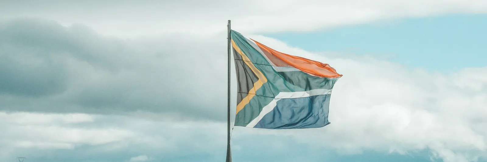 The South African flag flying from a pole against soft overcast sky, with dark mountain ridges along the lower third of the frame. Muted, restrained colour. Lan