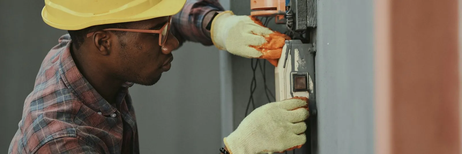 A Black African electrician in a yellow hard hat and work gloves wiring an electrical box on a wall, shot in profile. Landscape, 3-2.