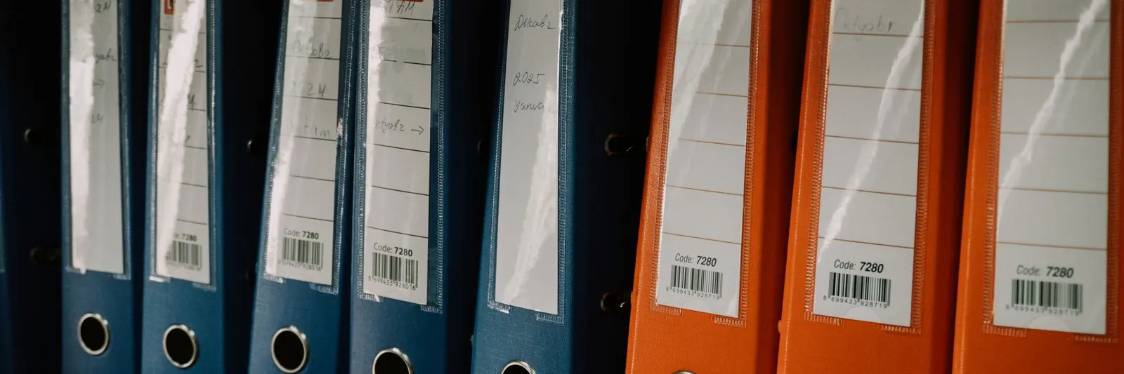 A row of blue and orange lever-arch binders with handwritten spine labels standing on a records-room shelf, side light raking across them. Landscape, 1600x1067.