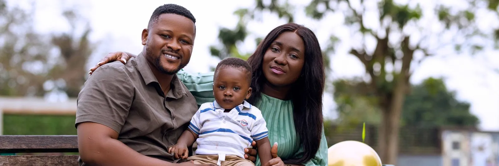 Two parents seated on a park bench holding their young toddler son, shot outdoors in natural light in Kumasi, Ghana. Landscape 1600x1067.