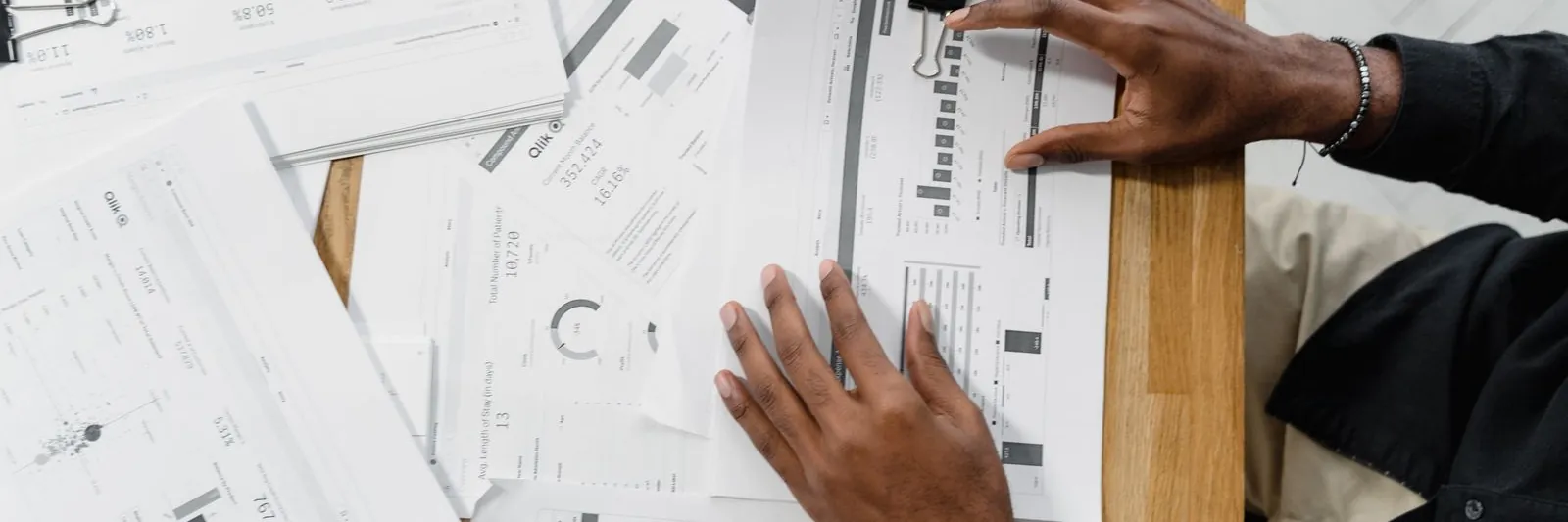Overhead shot of a person's hands sorting and comparing financial statements spread across a wooden desk, with a phone at the edge of the frame. Landscape 1600x