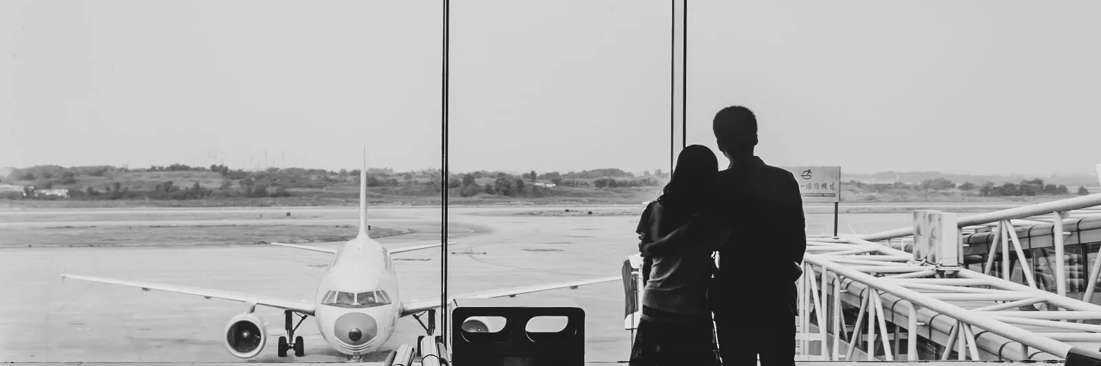 Black-and-white photograph of a couple standing together with an arm around each other, seen from behind, looking out through an airport terminal window at a pa