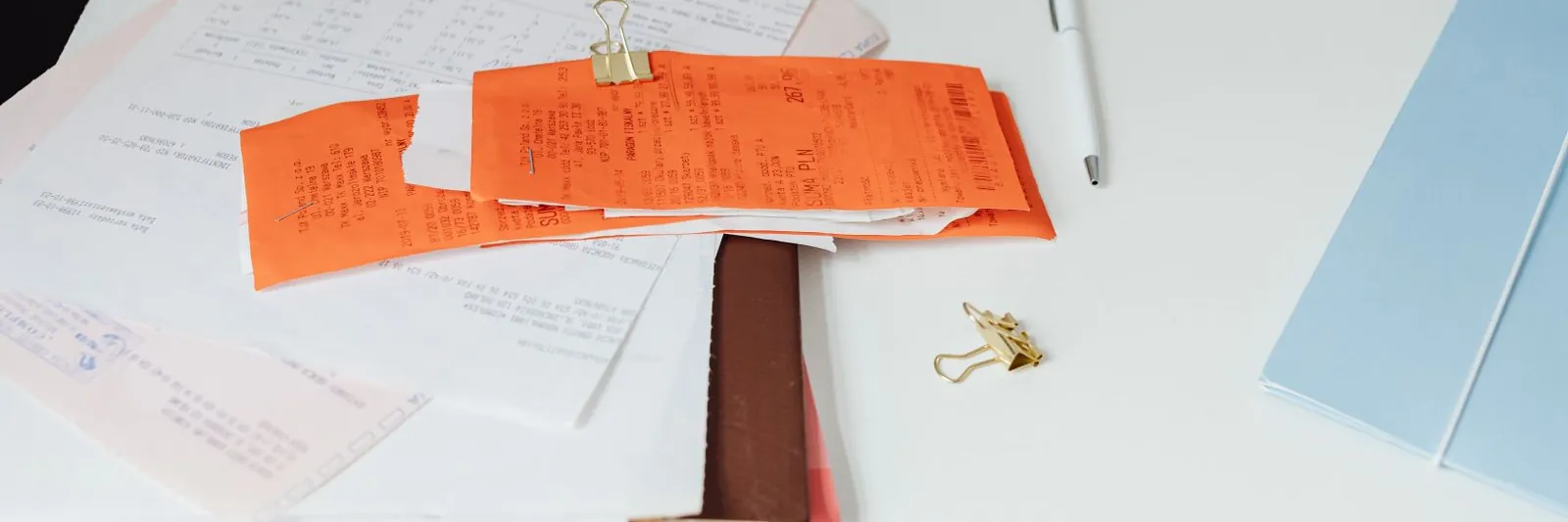 Overhead shot of a white desk with a stack of till slips and receipts clipped together, printed statements, binder clips, a pen and pencil, sticky notes and an