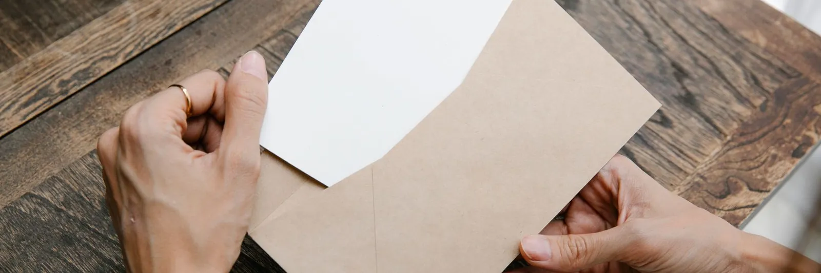 Overhead close-up of two hands drawing a blank white letter out of a kraft paper envelope on a dark wooden table, lit by soft window light.