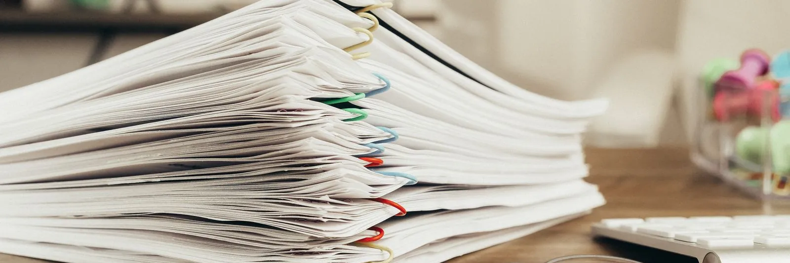 A thick stack of paper-clipped printed documents on a wooden desk, reading glasses resting in front, warm daylight, keyboard edge visible at right. No people, n